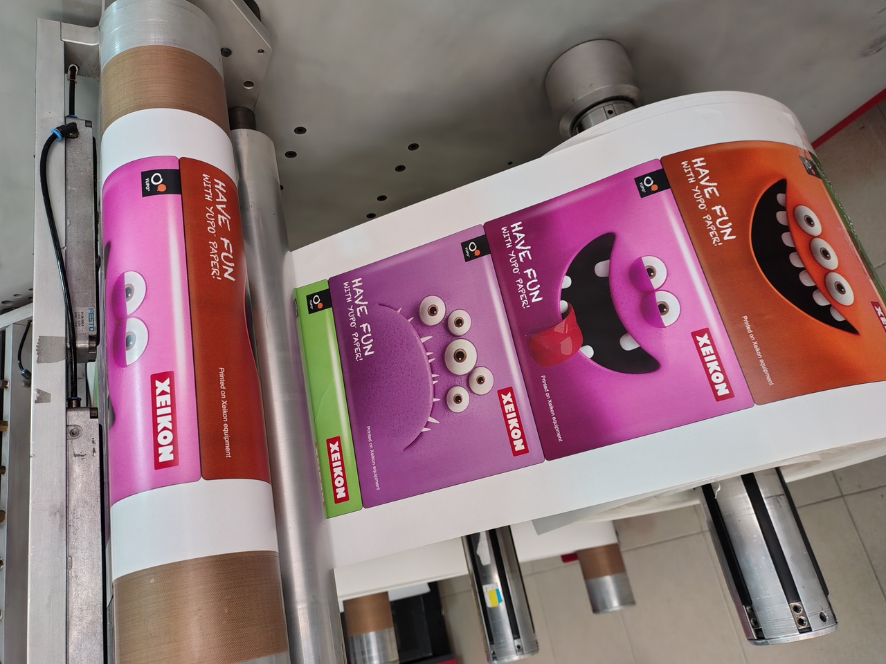 Impresora industrial de etiquetas imprimiendo diseños a color con rollos de papel y etiquetas adhesivas sobre una mesa blanca Impresora industrial de etiquetas imprimiendo diseños a color con rollos de papel y etiquetas adhesivas sobre una mesa blanca