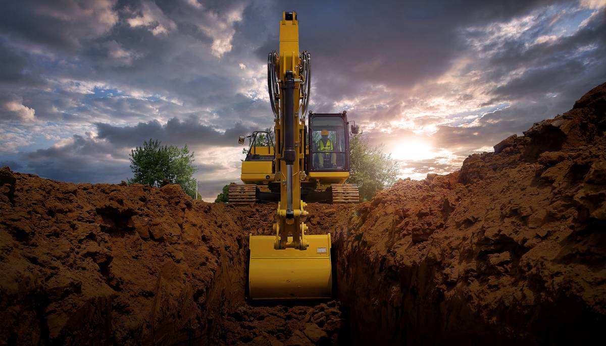 Excavadora amarilla operando en un sitio de construcción al atardecer, con cielo nublado y árboles al fondo.