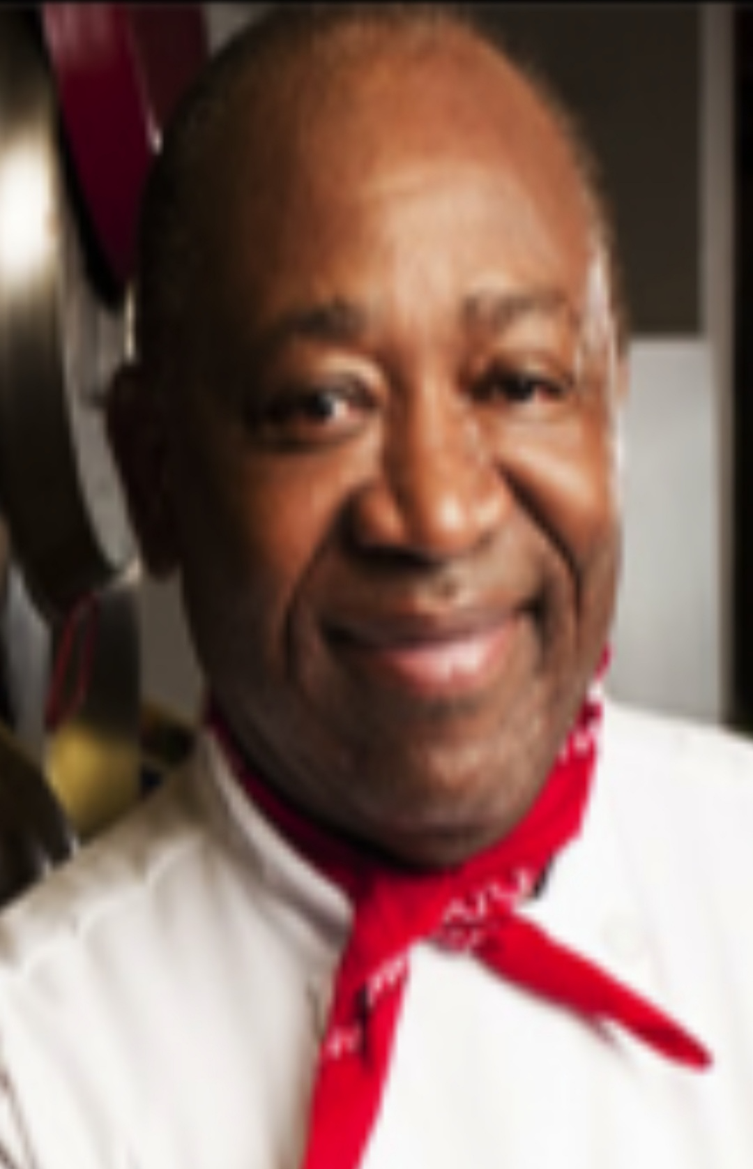 Smiling chef wearing a white uniform with a red neckerchief in a kitchen setting.