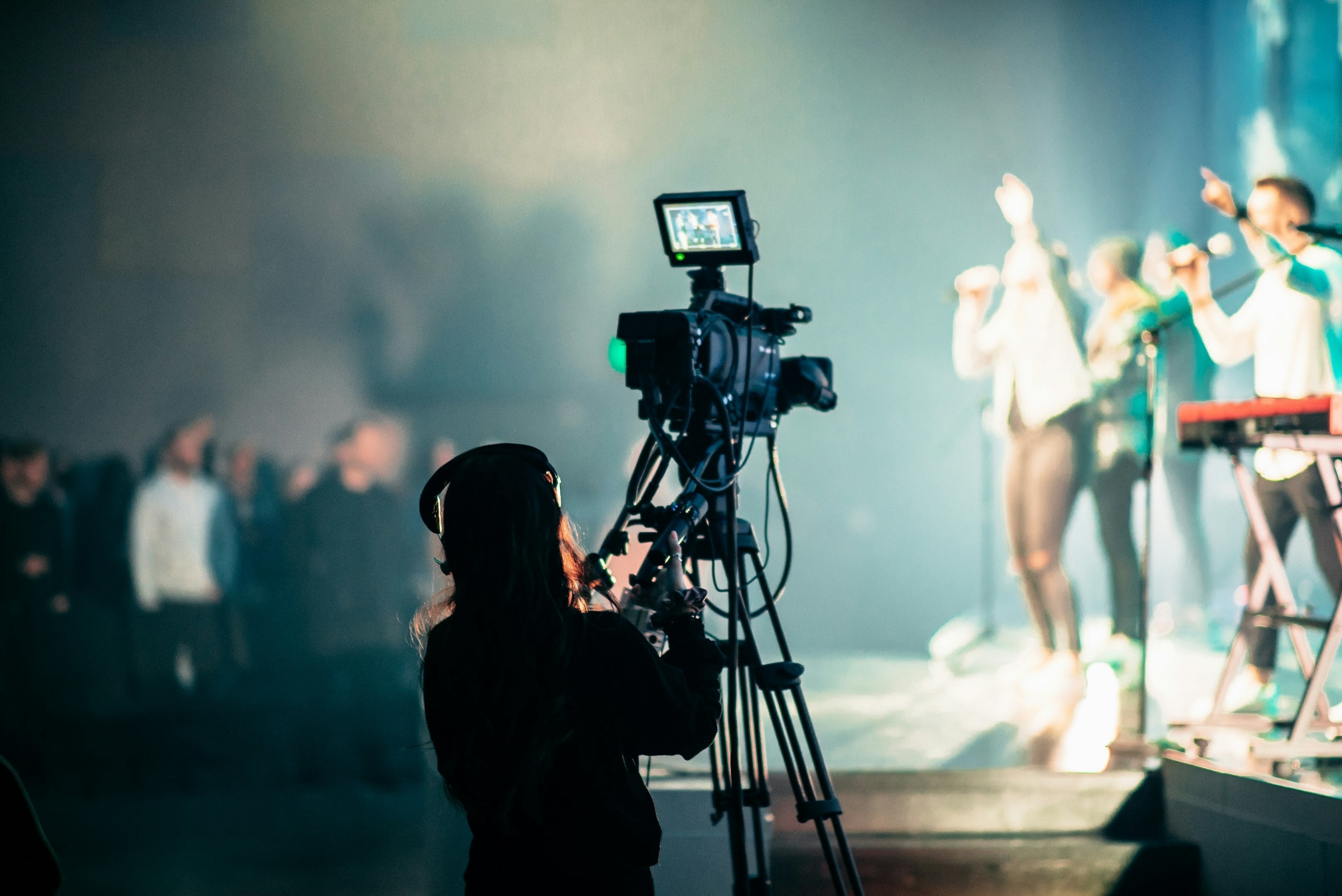 Cámara profesional grabando un concierto en vivo con músicos desenfocados en el escenario iluminado
