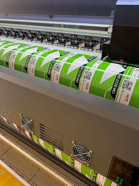 Impresora industrial imprimiendo etiquetas verdes con códigos QR y barras en un taller de impresión.