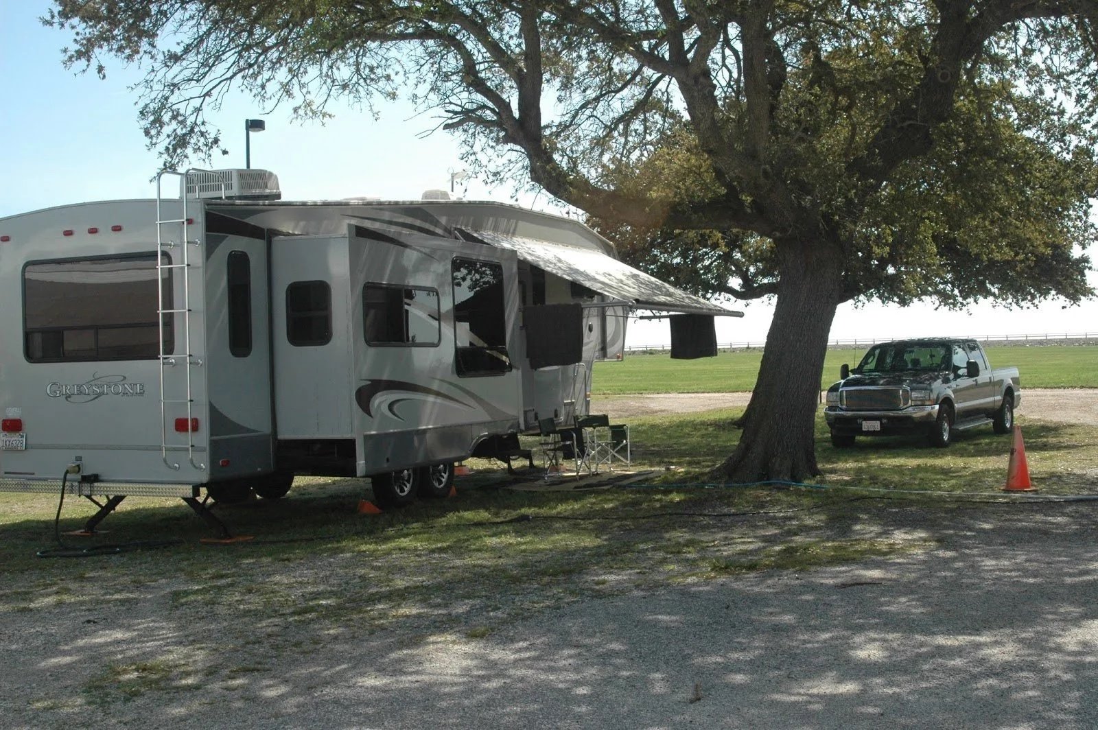 Travel trailer parked under a tree near a pickup truck on a sunny day at a campsite.