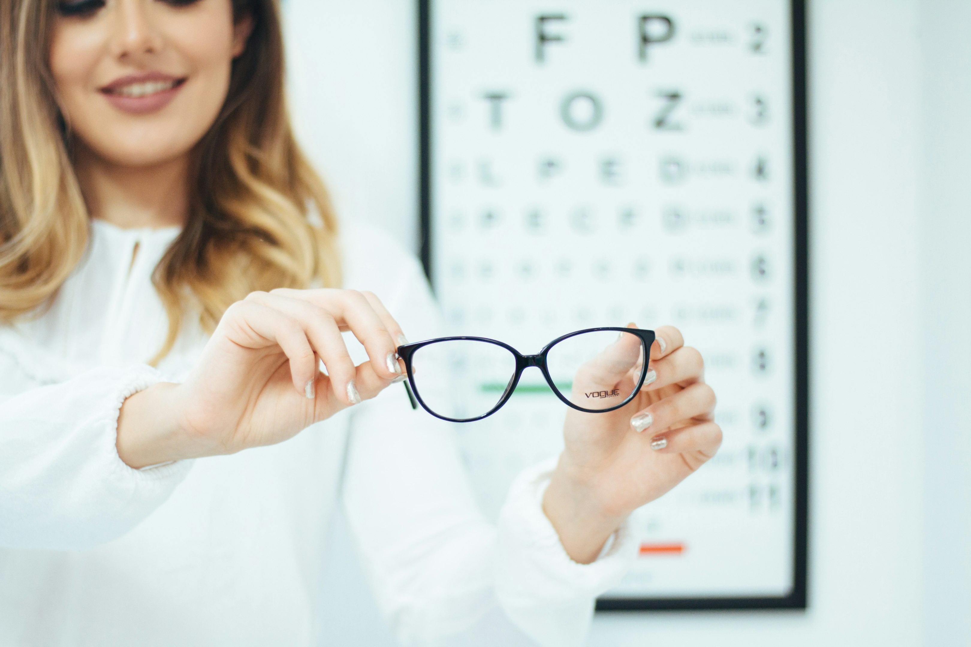 Mujer sosteniendo gafas delante de un cuadro de agudeza visual en una consulta de optometría.