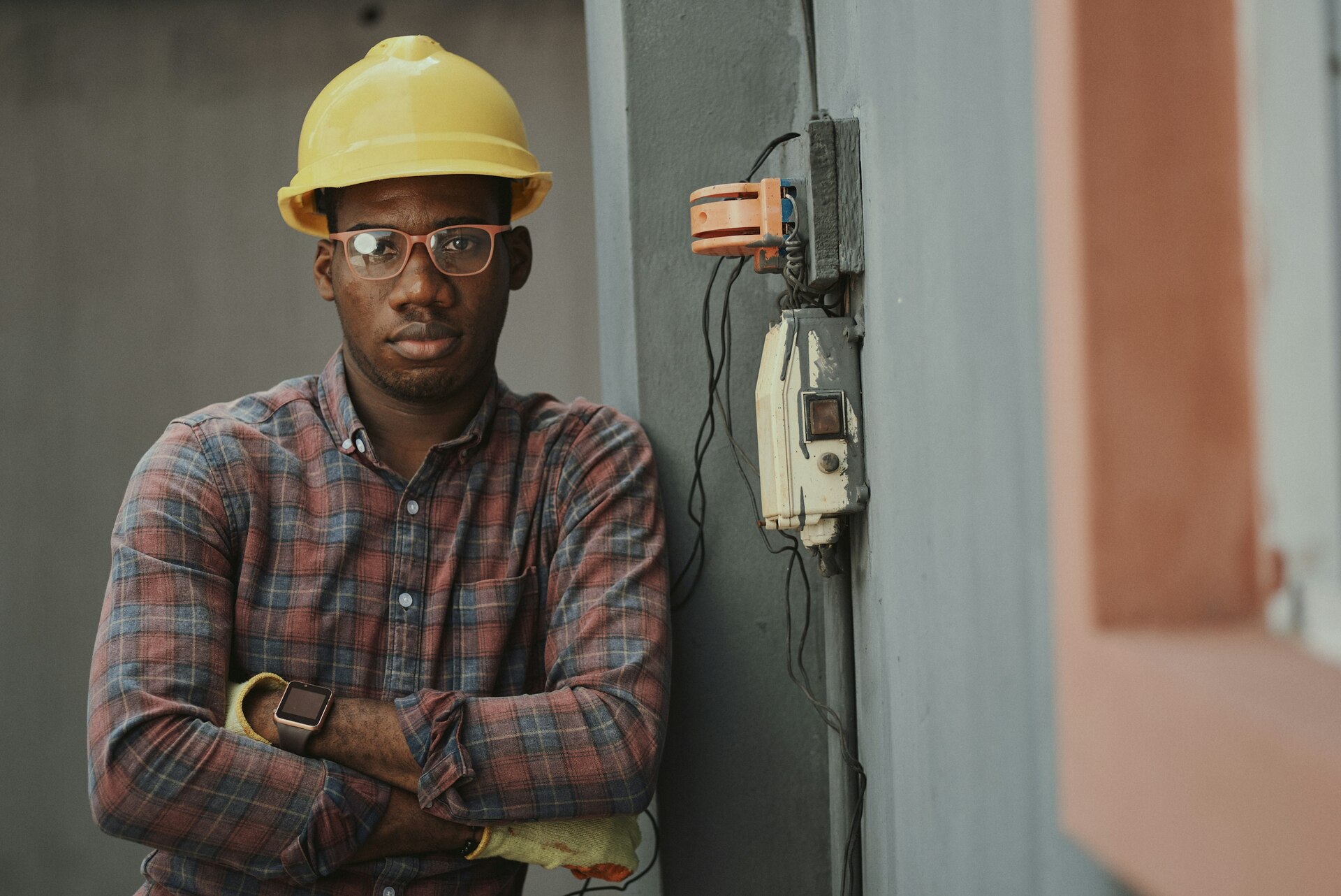 Trabajador con casco amarillo y gafas frente a una pared con cables y un dispositivo eléctrico. Trabajador con casco amarillo y gafas frente a una pared con cables y un dispositivo eléctrico.