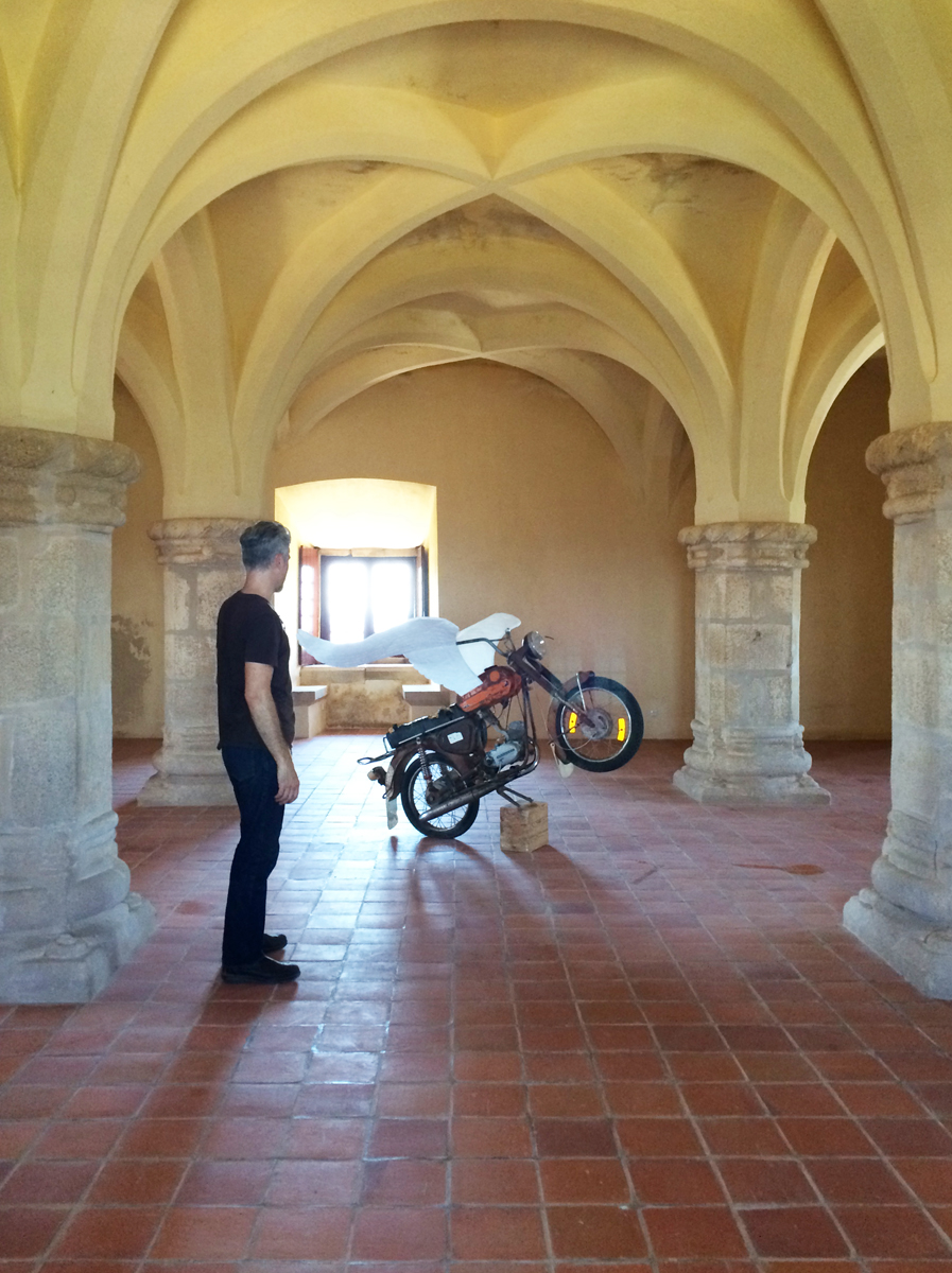 A man and a winged motorcycle in a room with stone pillars and vaulted ceiling.