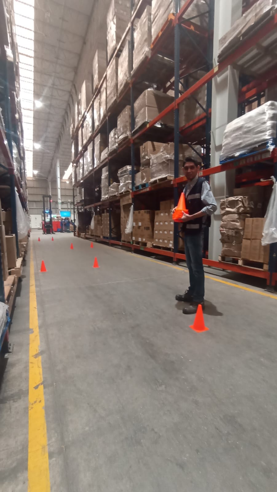 Hombre sosteniendo conos de seguridad en un almacén con estantes llenos de paquetes y luces en el techo.