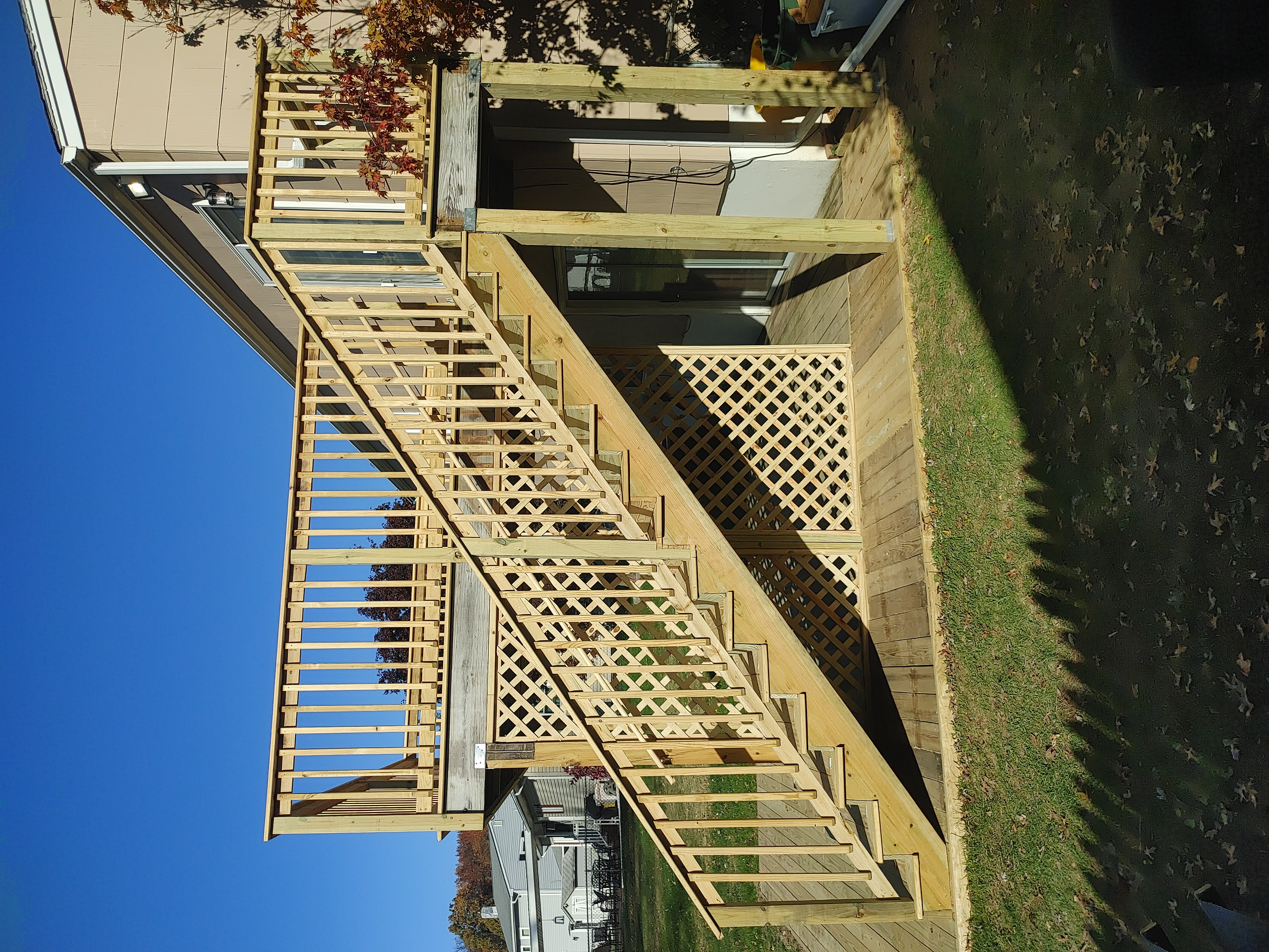 Two-level wooden deck and stairs attached to a house on a sunny day, featuring lattice panels and railings.