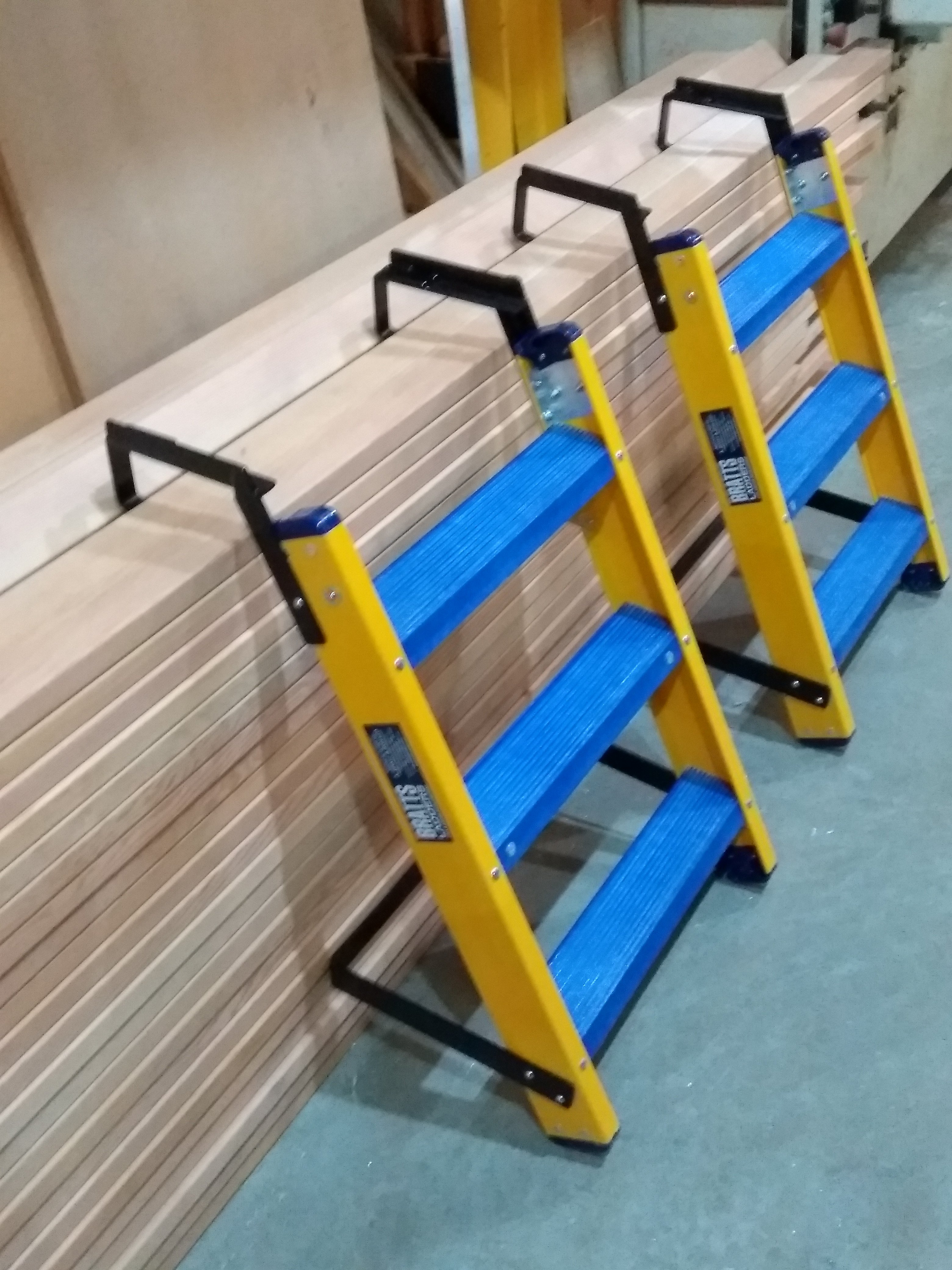 Three yellow and blue step ladders leaning against a stack of wooden planks in a workshop.