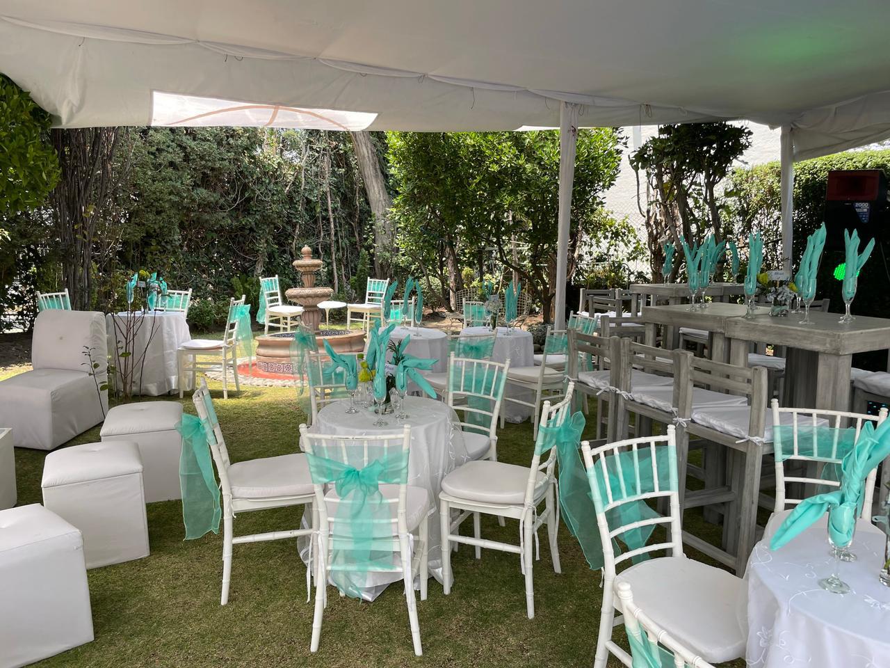 Decoración de boda al aire libre, con sillas blancas decoradas con tela azul turquesa y mesas junto a una fuente en un entorno de jardín. Decoración de boda al aire libre, con sillas blancas decoradas con tela azul turquesa y mesas junto a una fuente en un entorno de jardín.