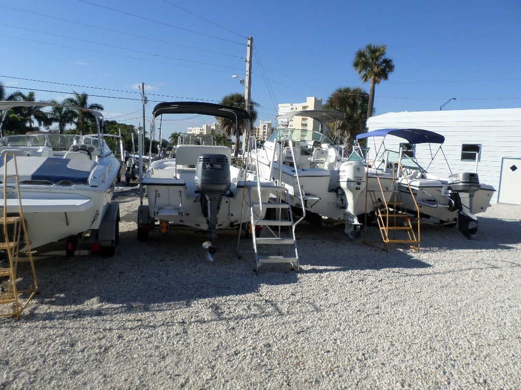 Boats for sale lined up on a gravel lot with outboard motors, ladders, palm trees, and clear blue sky