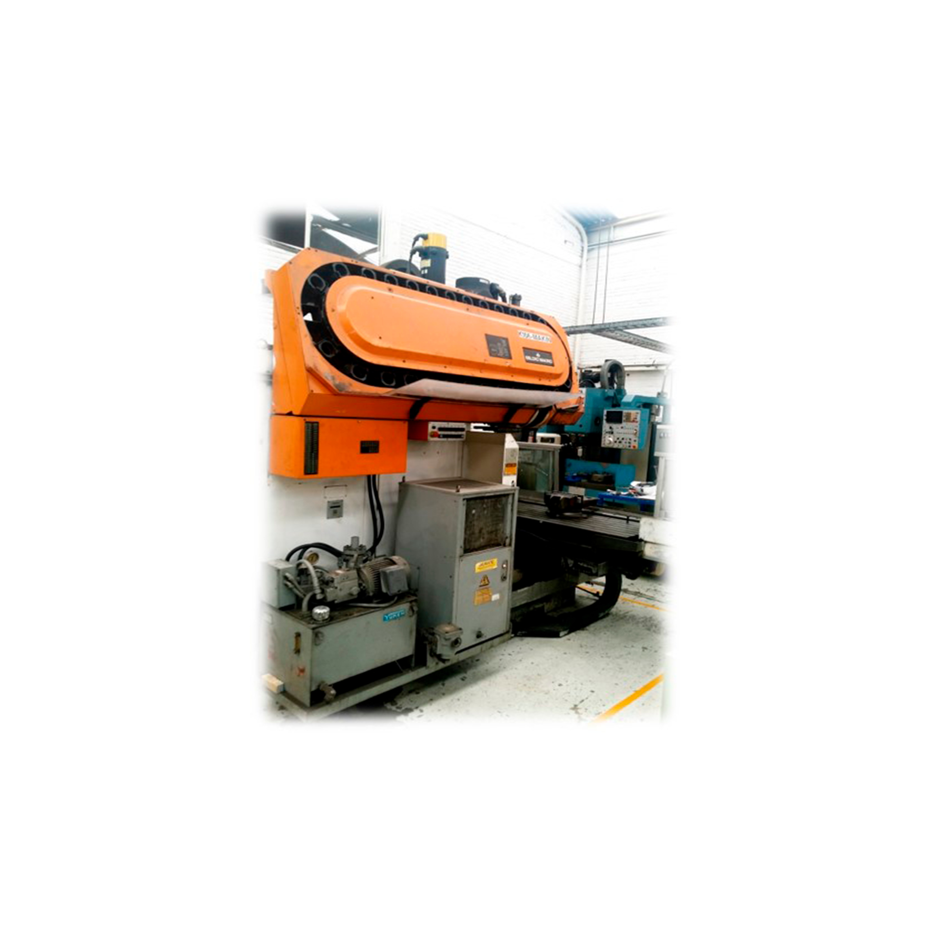 Máquina industrial de color naranja en un taller, con equipo de control y herramientas alrededor.