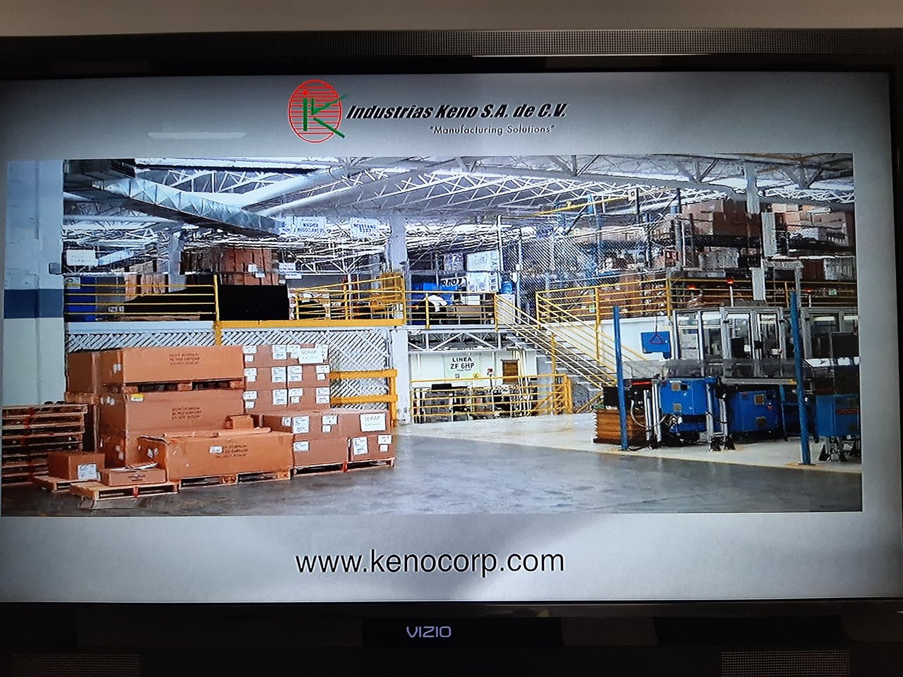 Interior de fábrica con cajas apiladas, maquinaria y estructura metálica en el techo.