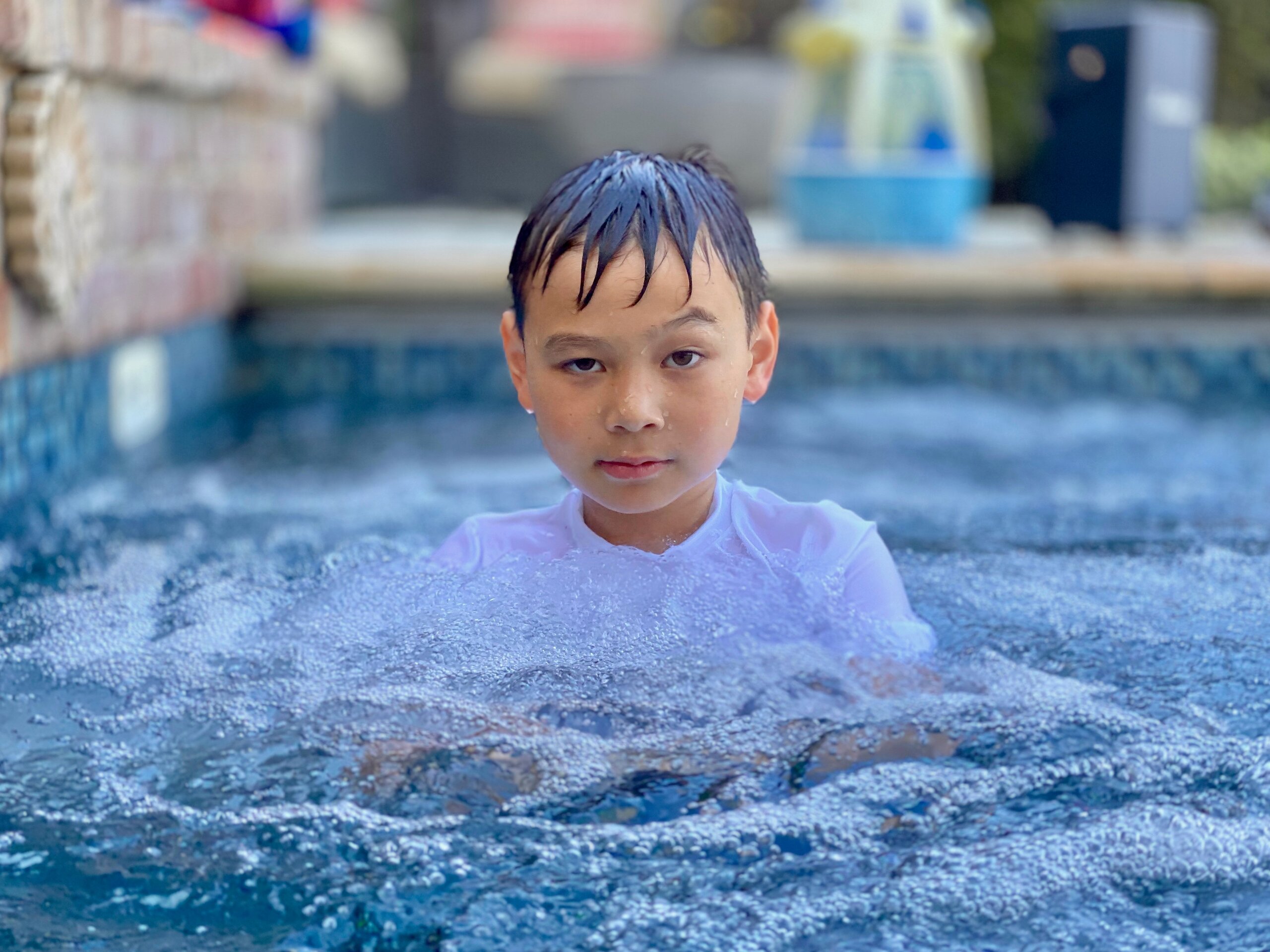 Young boy in a swimming pool with wet hair and a white shirt, surrounded by bubbling water.