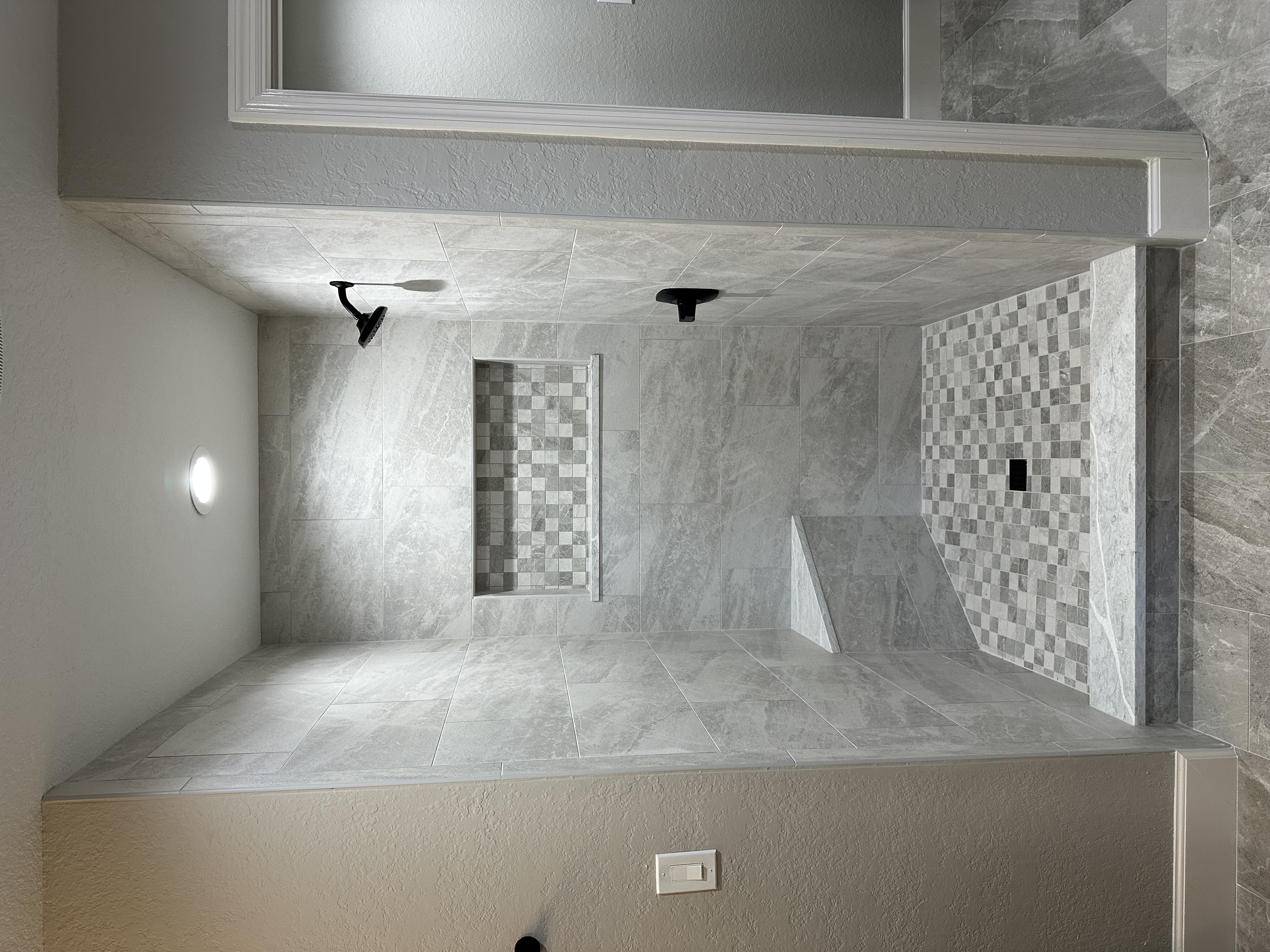 A spacious walk-in shower with 12x24 porcelain tiles, a roomy niche, corner bench, and a stylish mixed white and silver 2x2 shower floor, finished off with matte black fixtures for a modern touch.