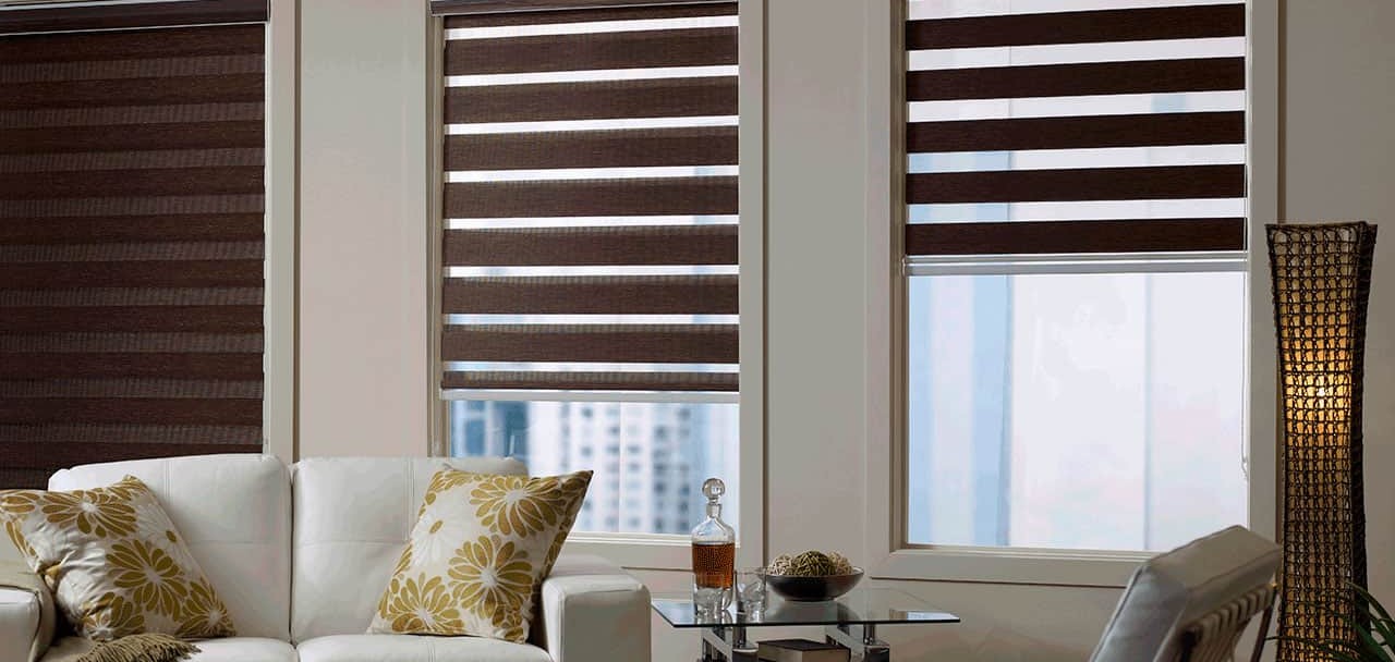 Modern living room with white sofa and floral cushions, featuring striped roller blinds on large windows.