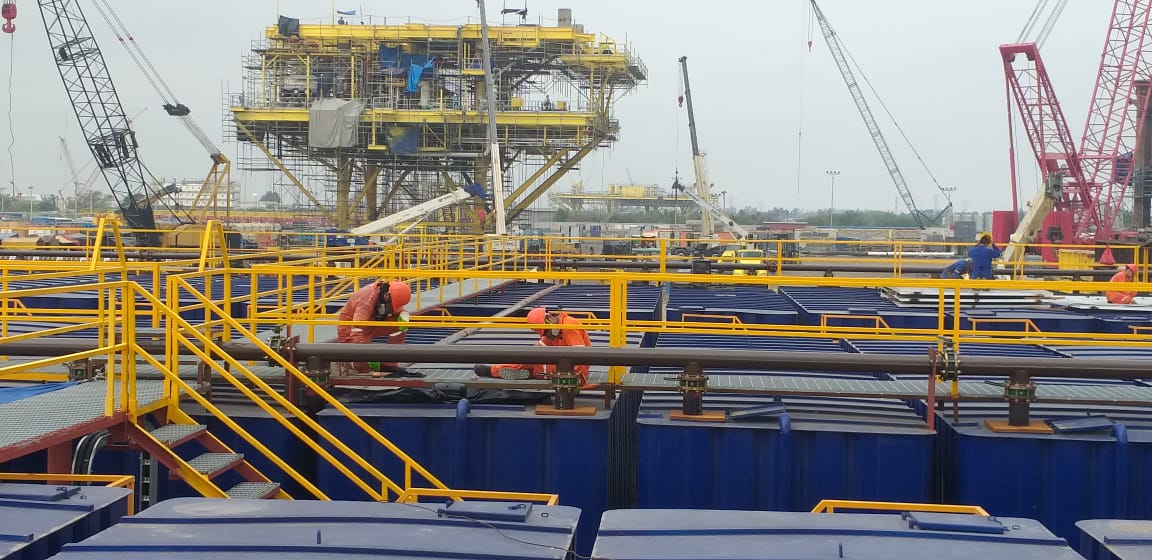 Construction workers on an industrial offshore rig with cranes and steel framework in the background.