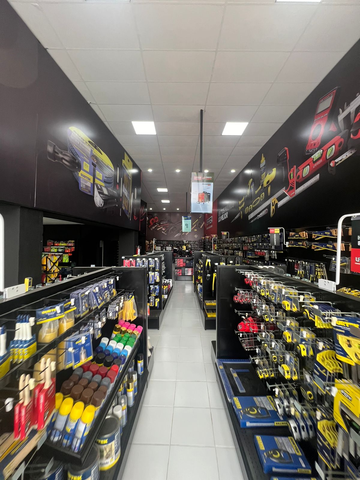 Pasillo de una tienda de herramientas con estantes llenos de productos como aerosoles, brochas y herramientas manuales, con carteles de marca en las paredes.