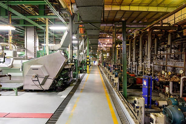 Interior de una planta industrial con maquinaria y tuberías alineadas a lo largo de un pasillo iluminado.