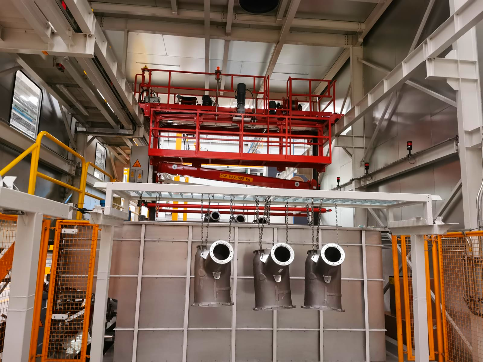 Instalación industrial con plataforma elevadora roja y piezas metálicas colgadas en línea de producción