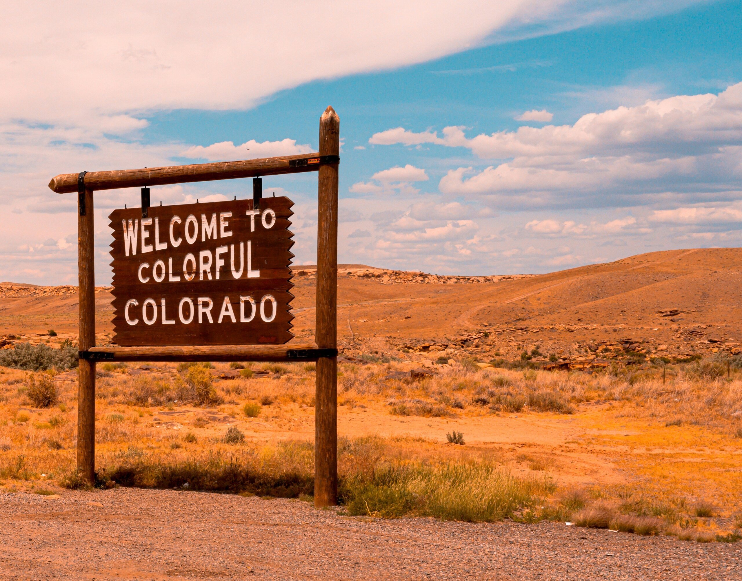 Welcome to Colorful Colorado sign in a desert landscape under a blue sky with clouds.
