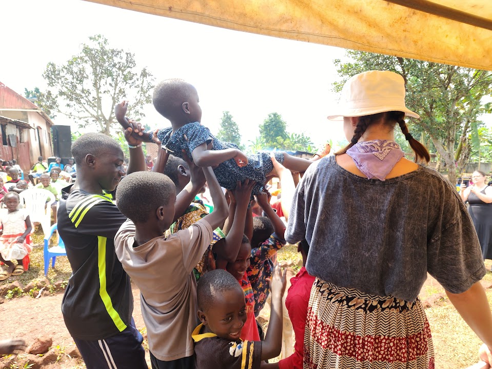 Children help lift a smaller child in a playful outdoor community gathering, with an adult woman wearing a hat nearby.