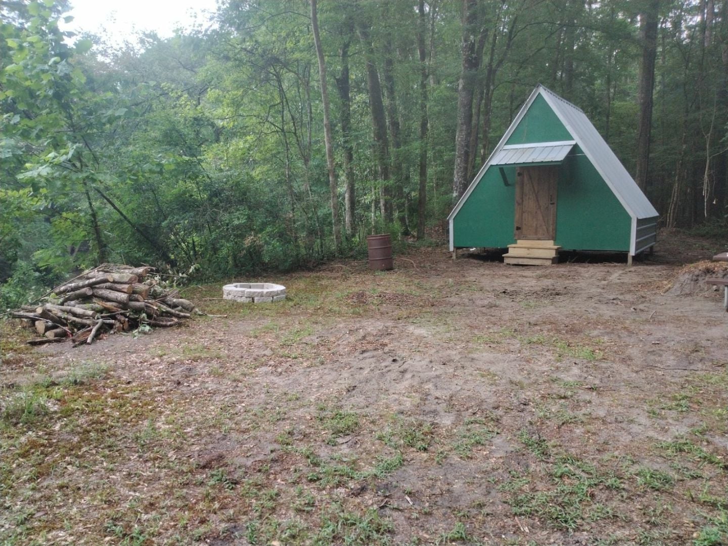 Rustic green A-frame cabin in a forest setting with stacked firewood and fire pit.