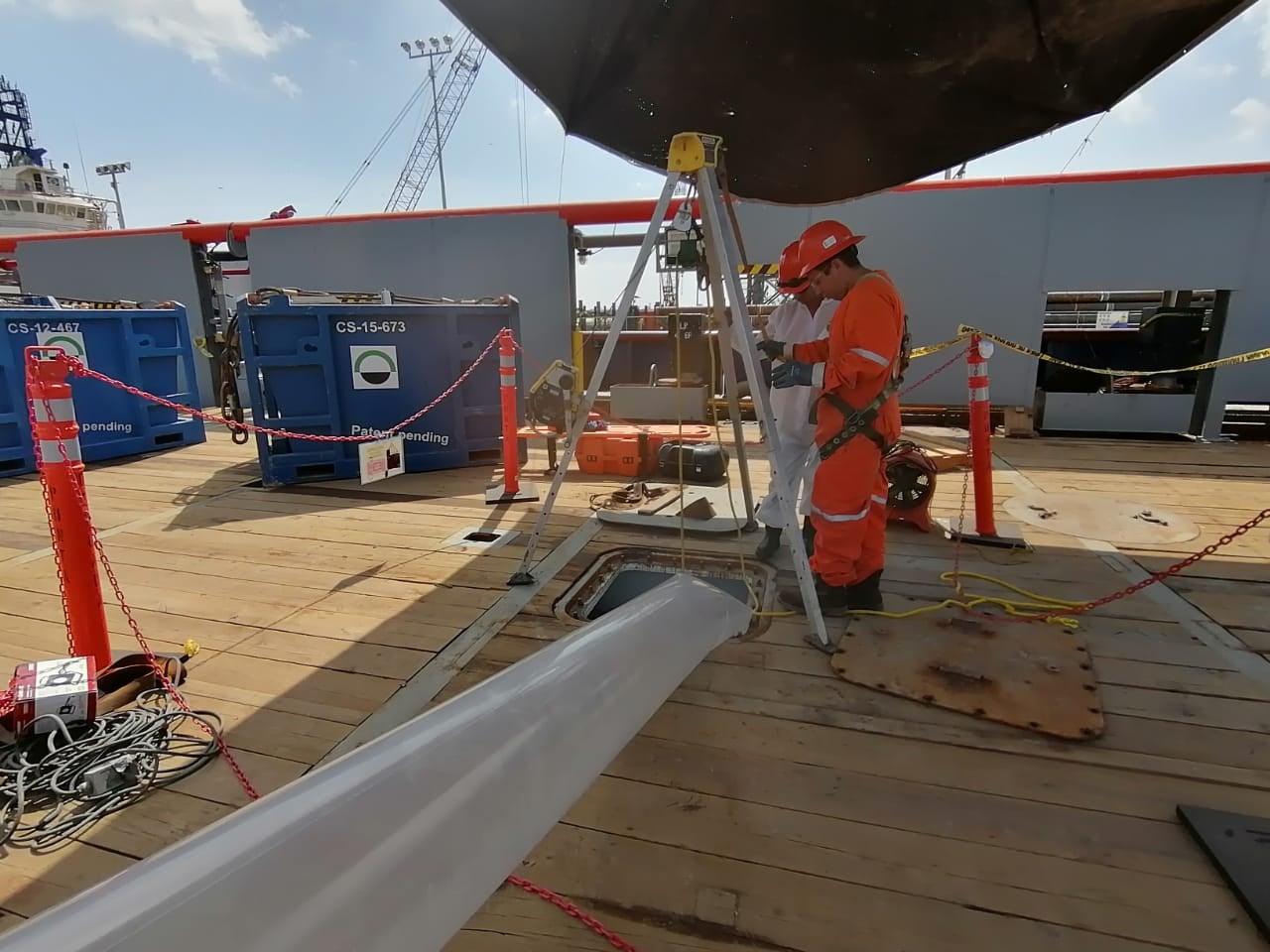 Two workers in safety gear operate machinery on a ship deck, with tools and equipment visible around them.