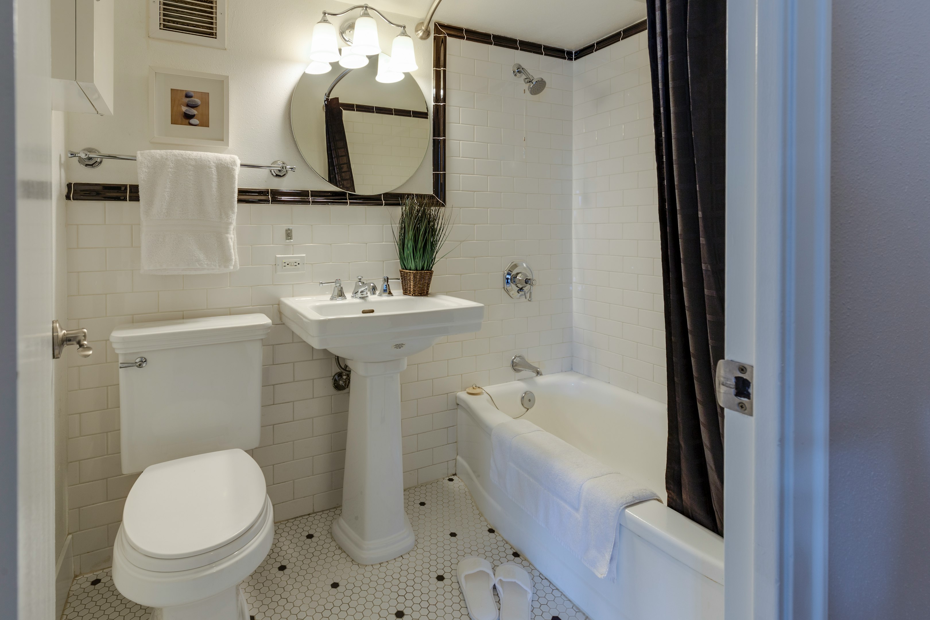 Modern white bathroom with pedestal sink, round mirror, subway tile, and bathtub with plant decor.