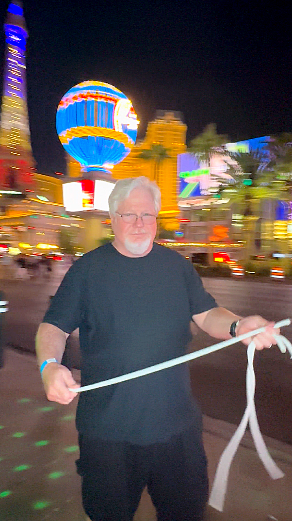 Man in black suit with surprised expression, holding playing cards tucked in his pocket against a plain background.