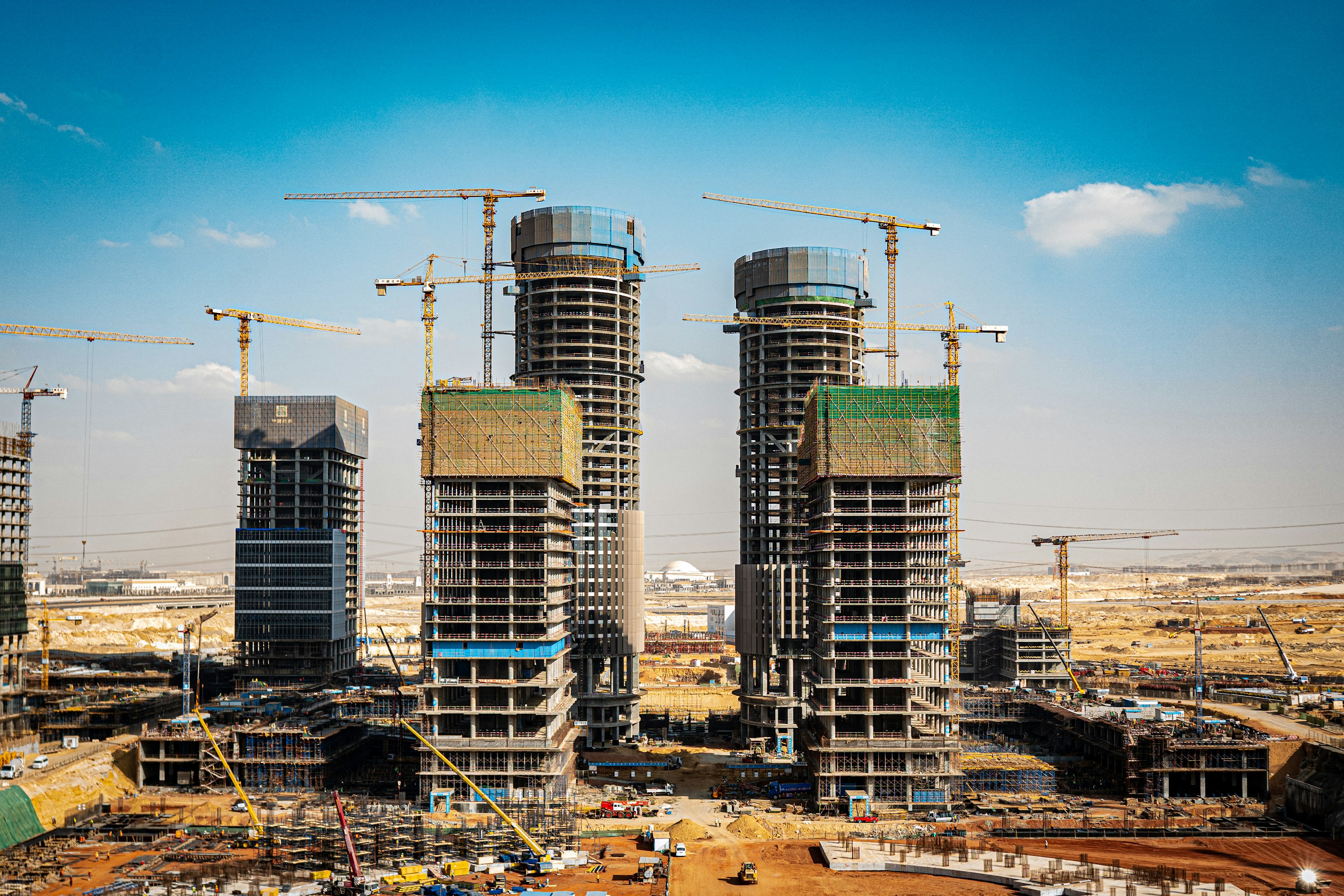 Panoramic view of a large construction site with multiple high-rise buildings and cranes against a clear blue sky.