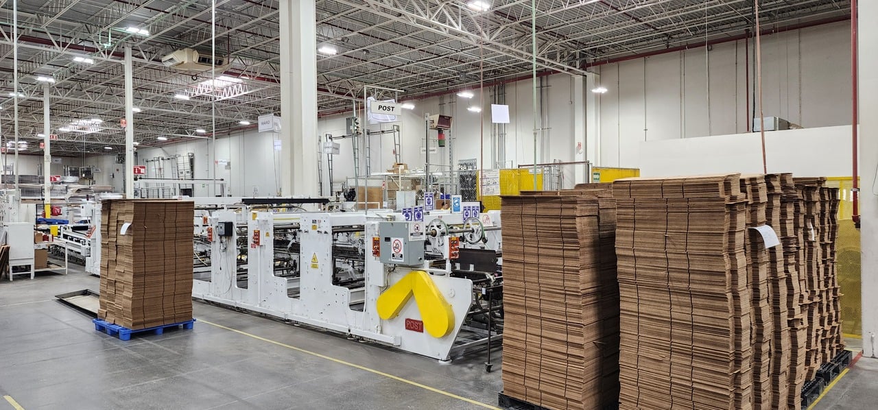 Fábrica de cajas de cartón con pilas apiladas junto a maquinaria industrial en un ambiente bien iluminado.
