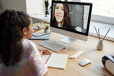 Girl participating in an online class on a desktop computer with a notebook and pencil on a wooden desk.