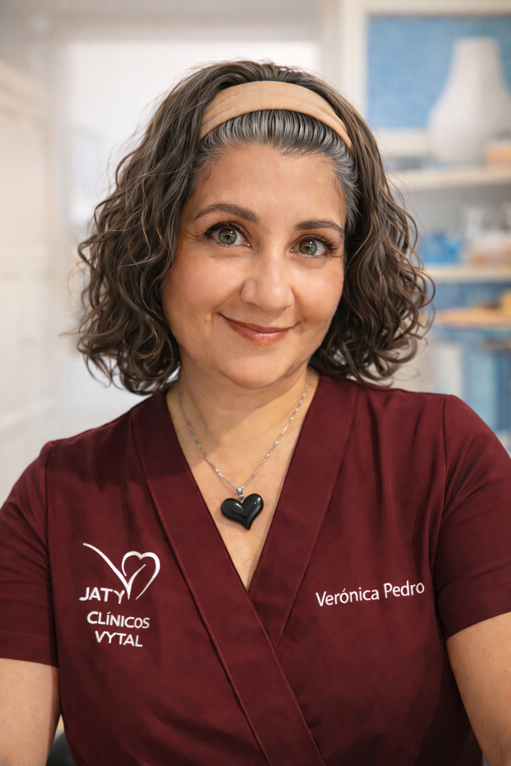 Mujer con uniforme de médico color burdeos, con cabello rizado corto y un collar de corazón negro, en un entorno profesional.