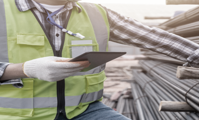 Trabajador de construcción con chaleco reflectante y guantes usando una tableta en un sitio con barras de acero. Trabajador de construcción con chaleco reflectante y guantes usando una tableta en un sitio con barras de acero.
