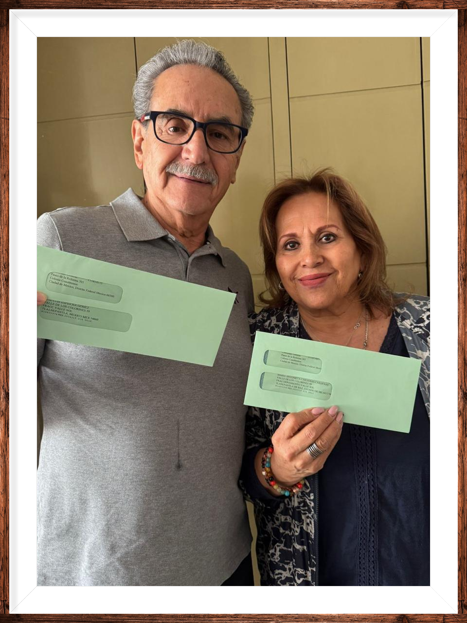 Pareja adulta sonriendo y sosteniendo sobres verdes frente a un fondo de armario beige.