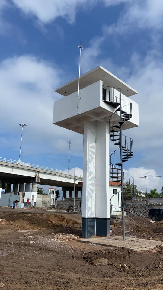 Torre de vigilancia blanca con escalera de caracol cerca de un puente en construcción, bajo un cielo azul. Torre de vigilancia blanca con escalera de caracol cerca de un puente en construcción, bajo un cielo azul.