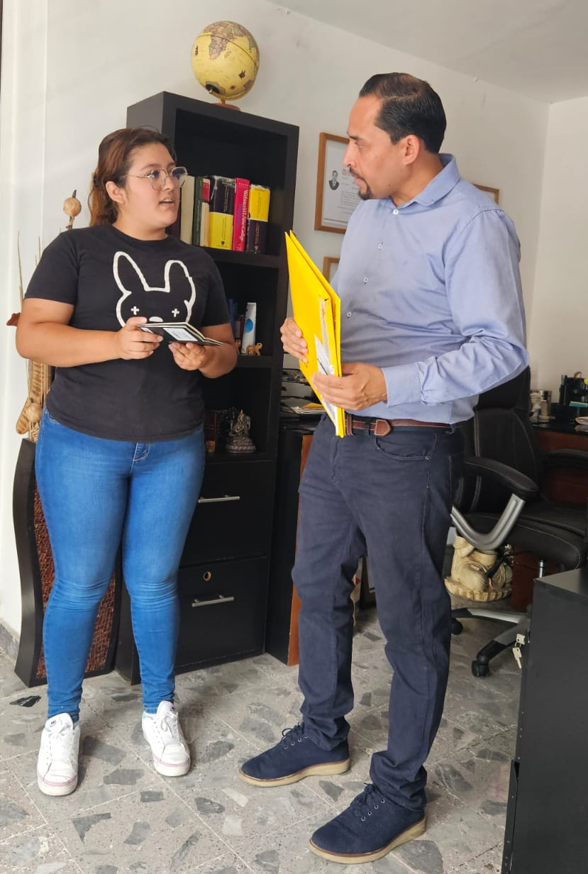Dos personas conversando en una oficina, una mujer sostiene un dispositivo y un hombre con camisa azul lleva carpetas amarillas, estante con libros y un globo terráqueo detrás.
