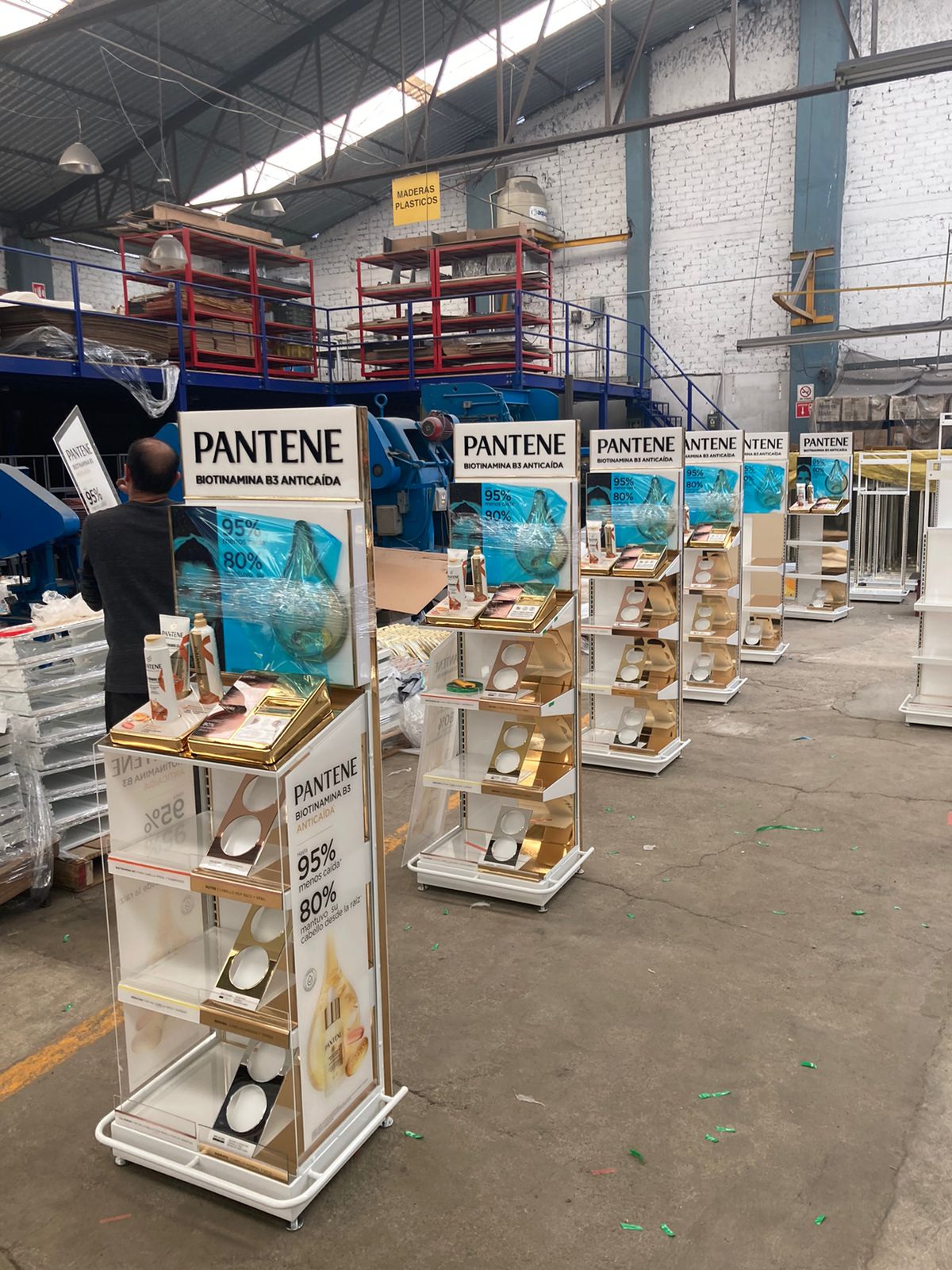 Exhibidores de productos Pantene dentro de una fábrica con estructuras metálicas y estantes de madera en el fondo.