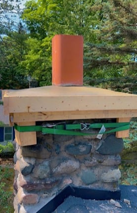 Stone chimney with a wooden and metal strap cap, surrounded by trees.