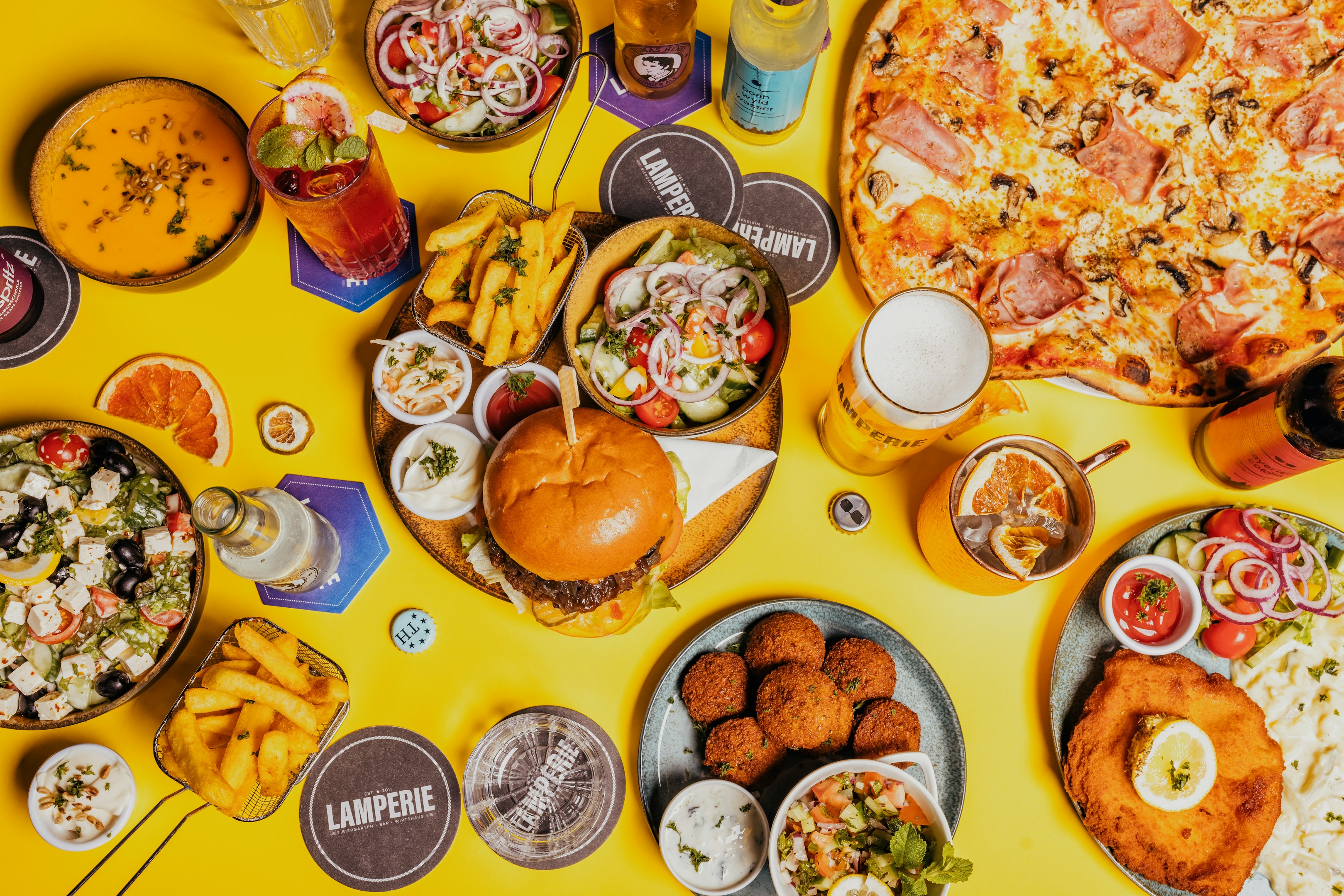 Colorful spread of a hamburger, pizza, falafel, salads, fries, sauces, and drinks including cocktails and beer on a vibrant yellow table.