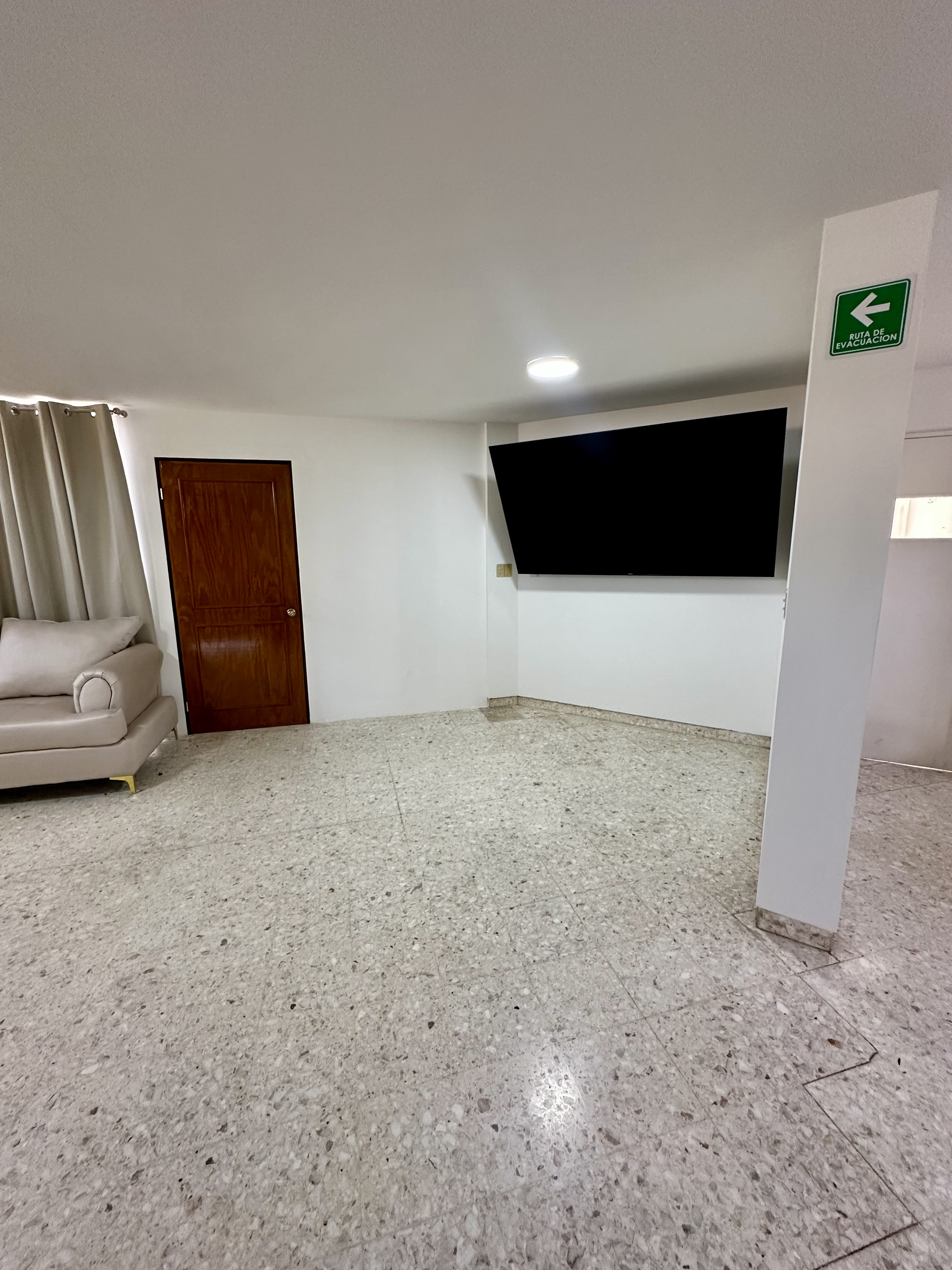 Mujer mayor sonriendo con gafas y chaqueta amarilla, sentada en sala de estar bien iluminada con plantas.