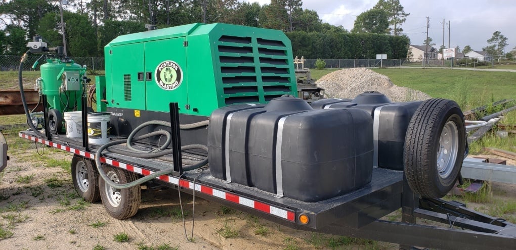 Mobile dustless blasting unit on a trailer with green machinery and black water tanks, surrounded by greenery and industrial background.