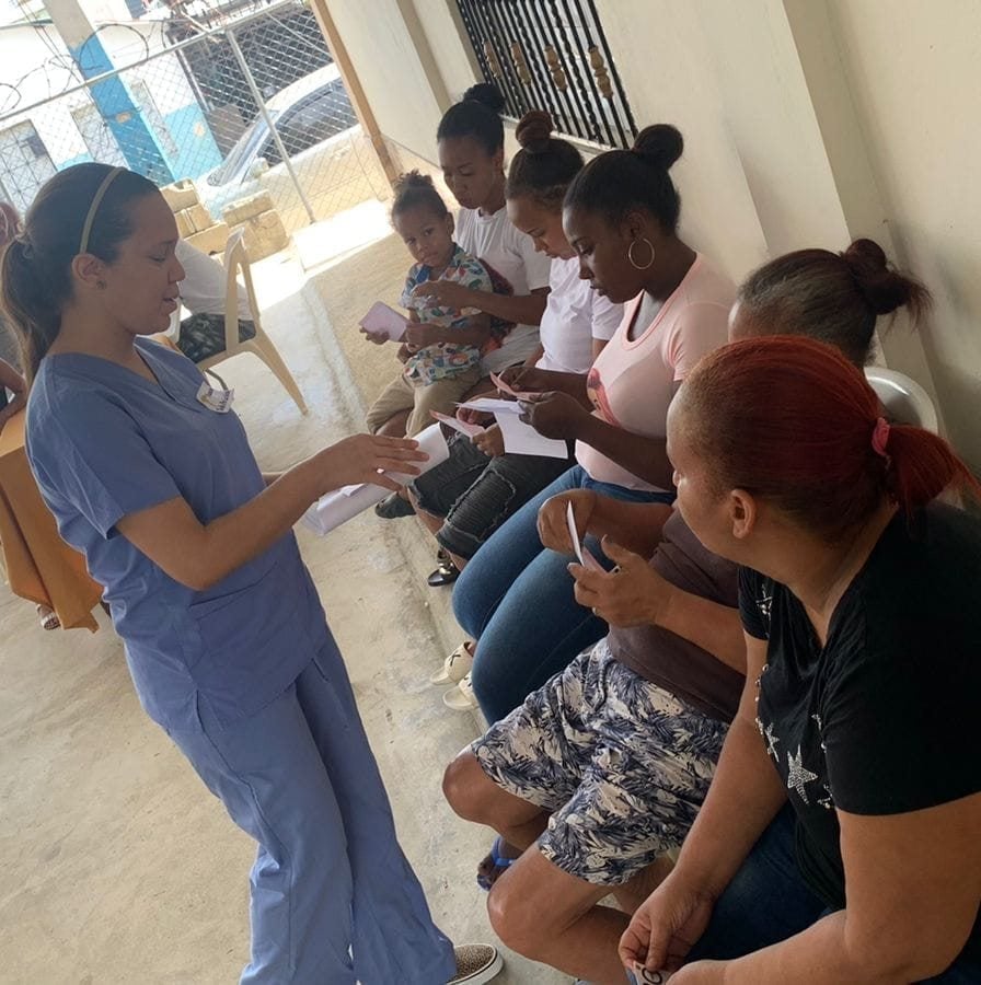 Mujer en uniforme médico entregando folletos a un grupo de personas sentadas en fila en un edificio, interacción comunitaria.