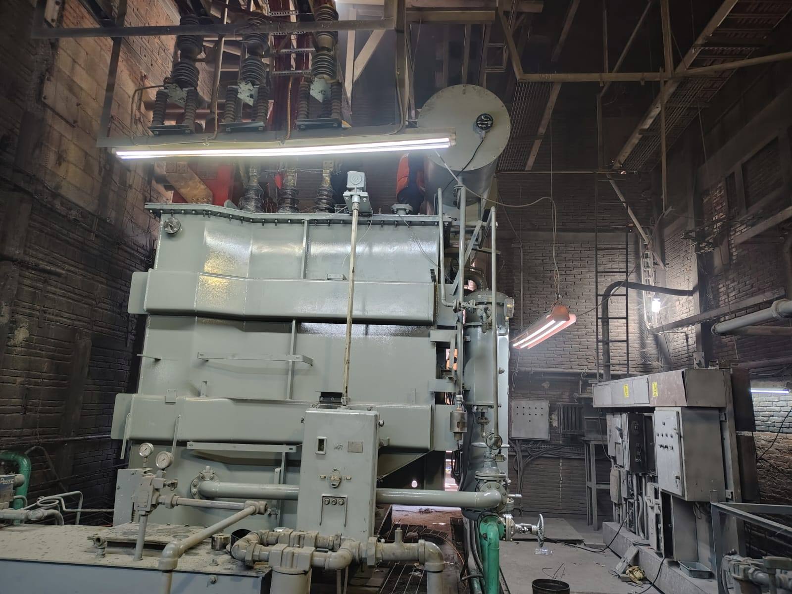 Hombre con uniforme de trabajo y casco en una fábrica moderna iluminada, rodeado de estanterías y equipos industriales.