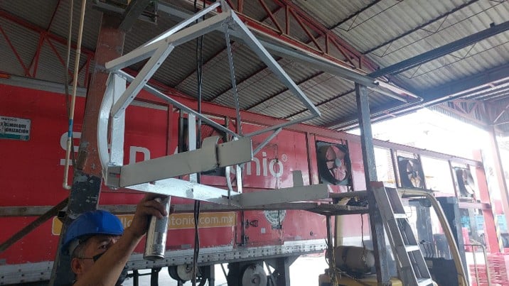 Un trabajador con casco azul realizando mantenimiento a una estructura metálica colgante frente a un camión rojo en un almacén.