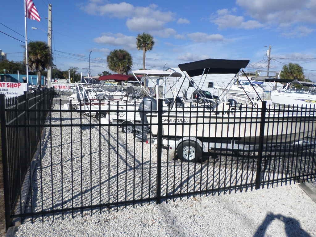 Boat dealership lot with trailered center console boats behind black metal fence under blue sky