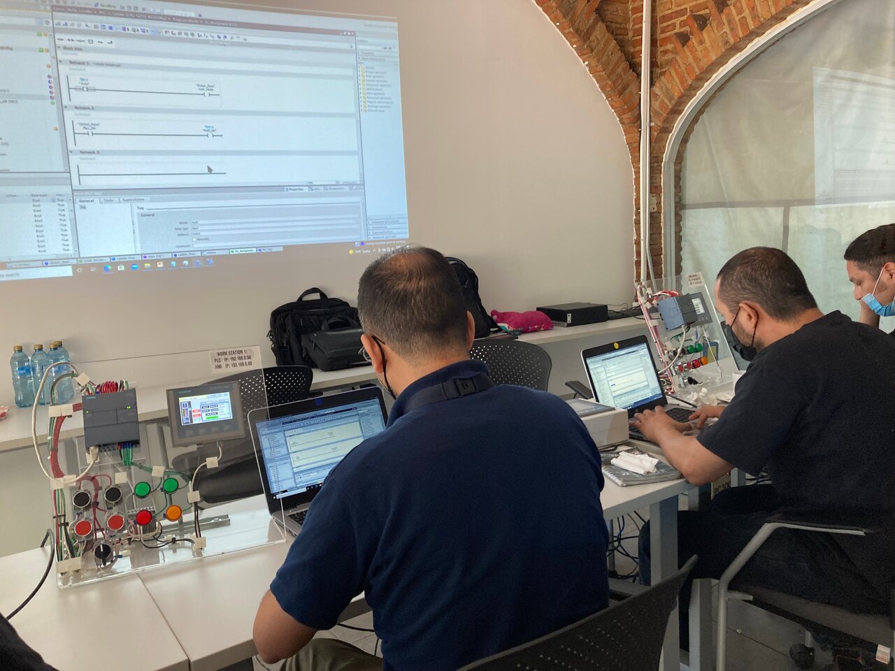 Personas trabajando en una sala de capacitación de programación PLC, con laptops y un sistema de control automatizado. Personas trabajando en una sala de capacitación de programación PLC, con laptops y un sistema de control automatizado.