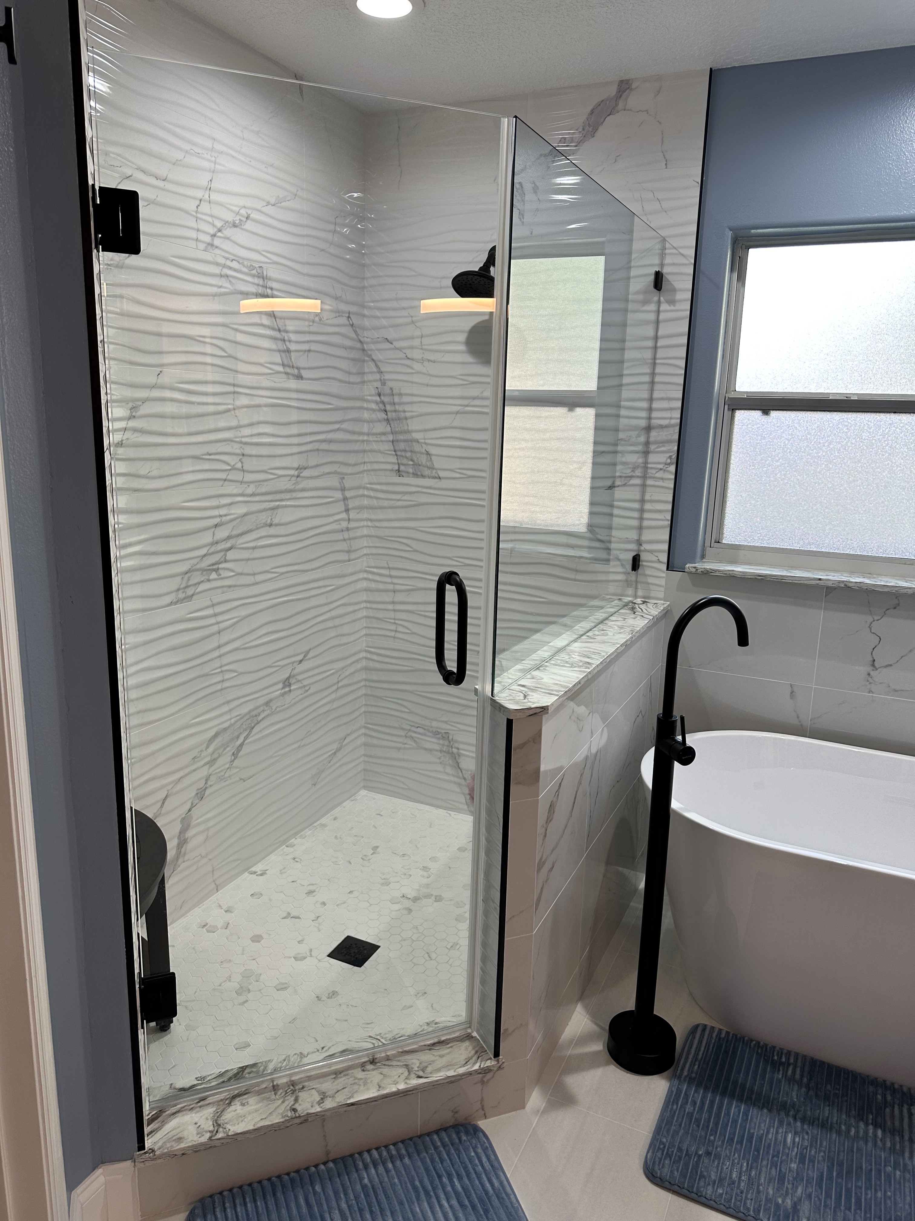 The 3/8" heavy clear glass door and panel with matte black hardware frame out this spacious walk-in shower showcasing the 12x24 Statuario glossy wave wall tile, matte hexagon shower floor, and black metal trim.