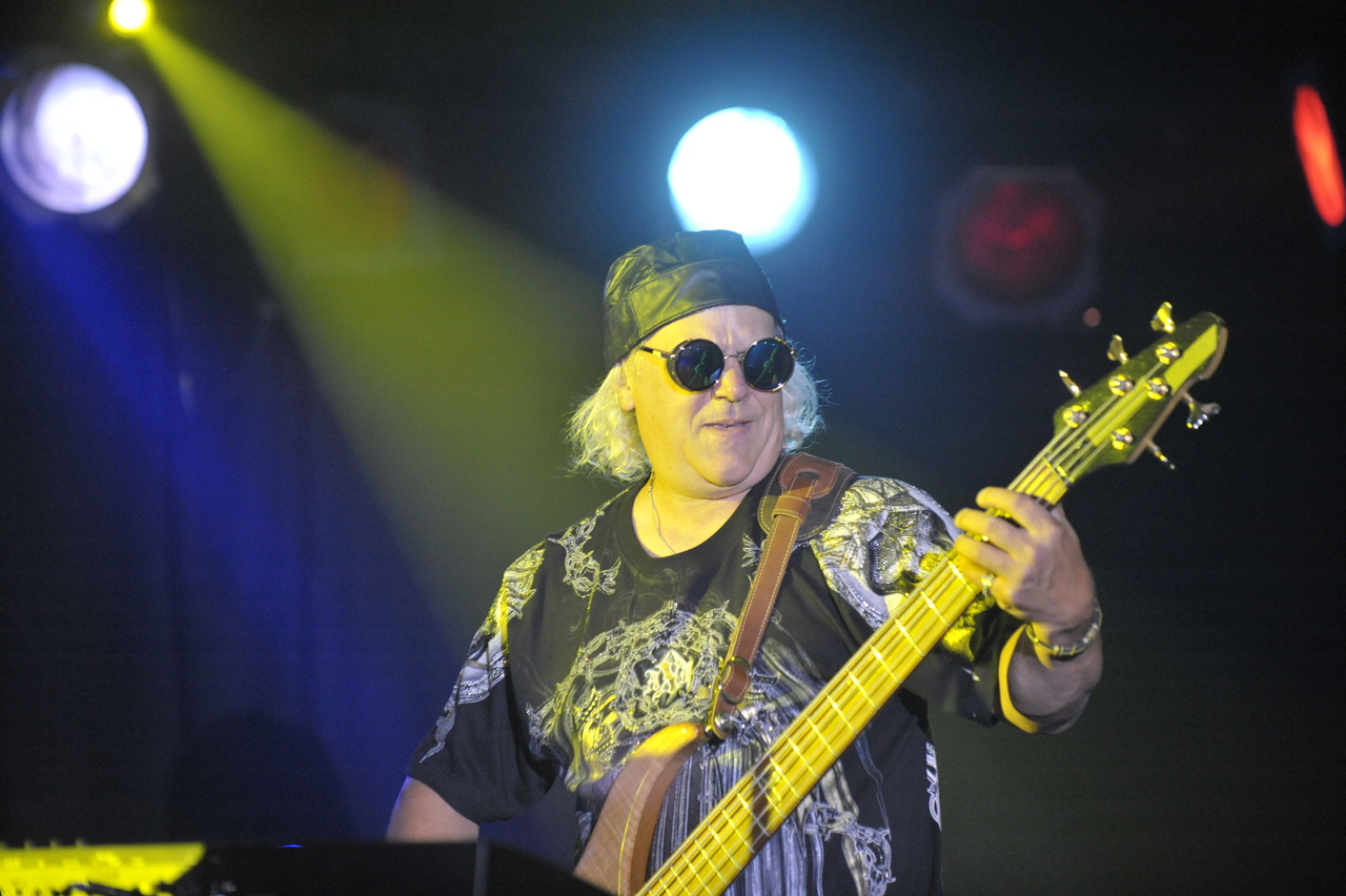Live band performing in a modern auditorium with a large audience, featuring guitarists, a drummer, and a double bassist, with artistic paintings and a map projection in the background.