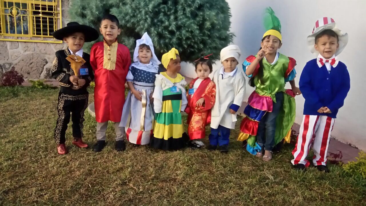 Niños pequeños sonriendo, vestidos con trajes típicos de diversos países, posando al aire libre frente a un arbusto.